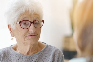 NIEUWS Een zorgverlener is in gesprek met een oudere vrouw die een grijs vest en een bril draagt.
